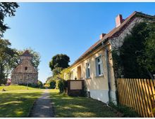 Ein gepflasterter Weg führt durch eine grüne Wiese zu einer kleinen steinernen Kirche mit rotem Dach. Rechts im Bild befindet sich ein gelbes Haus mit roten Dachziegeln, umgeben von Bäumen und einem Holzzaun.