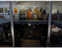 Innenansicht eines Museums oder Ausstellungsraums mit zwei Etagen. Auf der oberen Ebene sind bunte Schaufensterpuppen ausgestellt, während die untere Ebene mit einer dunklen Kulisse beleuchtet ist und verschiedene Figuren sowie hängende Objekte zeigt; die Architektur umfasst weiße Säulen und umlaufende Geländer.