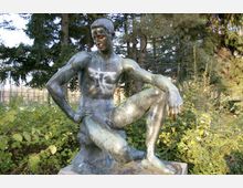 Bronzestatue eines sitzenden nackten Mannes in einer natürlichen Umgebung mit Bäumen und dichtem Gebüsch im Hintergrund. Die Statue zeigt erkennbar Graffiti auf der Oberfläche.