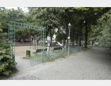 Städtischer Spielplatz mit Sandboden, umgeben von einem grünen Metallzaun und schattenspendenden Bäumen. Im Hintergrund sind Klettergerüste aus Holz, eine Rutsche und Sitzbänke zu sehen, neben einem gepflasterten Gehweg.