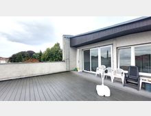 Geräumige Terrasse mit Holzboden und einer niedrigen Mauer, die einen Blick auf Bäume und Dächer bietet. An der Fassade befinden sich große Fenster und Türen sowie eine Leiter, daneben stehen mehrere weiße und schwarze Gartenstühle.