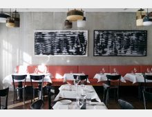 Innenraum eines modernen Restaurants mit eleganter Einrichtung. Es gibt rote gepolsterte Sitzbänke entlang der Wand, schwarz-weiße abstrakte Kunstwerke an der grauen Wand, schwarze Stühle und mit weißen Tischdecken gedeckte Tische, geschmückt mit Gläsern und Besteck.