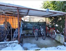 Überdachter Fahrradabstellplatz mit mehreren abgestellten Fahrrädern, umgeben von einem Metallgitterzaun. Der Boden ist teilweise mit Schnee bedeckt, im Hintergrund sind Büsche und eine orangefarbene Wand sichtbar.