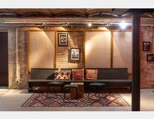 Gemütlicher Raum mit Backsteinwänden, einer langen Holzbank mit dekorativen Kissen in warmen Mustern, niedrigen Hockern, einem Holztisch und Teppichen mit bunten geometrischen Designs. Deckenrohre und warme Beleuchtung geben dem Raum einen industriellen Stil.