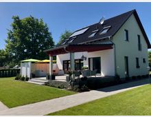 Moderne zweistöckige Einfamilienhaus mit weißer Fassade und schwarzem Satteldach, ausgestattet mit Solarpanelen. Eine großzügige, möblierte Terrasse mit bunten Sonnenschirmen grenzt an einen gepflegten Garten mit einem Rasen und umliegender Hecke, umgeben von Bäumen.