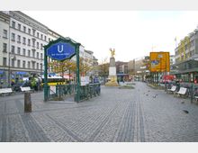 Stadtplatz mit gepflastertem Boden, im Zentrum ein U-Bahn-Eingang mit der Aufschrift „Bhf. Hermannplatz“, umgeben von historischen Gebäuden, Geschäften und Fußgängern. Im Hintergrund eine Goldstatue auf einem Sockel und Hinweisschilder für Verkehrsrouten.