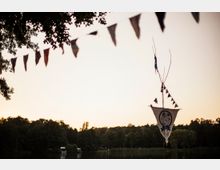 Fahnen sind an einem Ast und einer Schnur aufgehängt, davor ein Wimpel mit einem gemalten Symbol. Im Hintergrund ist ein See mit angrenzendem Wald bei Sonnenuntergang zu sehen.