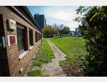 Backsteingebäude mit rechteckigen Fenstern und einem Schild mit der Aufschrift „Floris“ an der Wand, daneben ein gepflasterter Weg, der durch eine Wiese mit Bäumen führt. Im Hintergrund sind ein graues Hochhaus mit der Zahl „13“ sowie geparkte Fahrzeuge zu sehen.