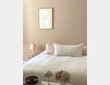 Minimalistisches Schlafzimmer mit einem hellen, beige gestrichenen Hintergrund, einem Bett mit weißer Bettdecke und einem großen weißen Zierkissen. Links steht ein kleiner Nachttisch mit einer cremefarbenen Lampe, an der Wand hängt ein abstraktes Bild, und im Vordergrund befinden sich ein kleiner Tisch mit einer Vase getrockneter Blumen und einer Tischlampe.