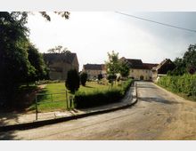 Eine ruhige ländliche Straßenszene mit gepflastertem Gehweg und Häusern im Hintergrund. Auf der linken Seite befindet sich eine kleine Grünfläche mit Bäumen und Hecken.