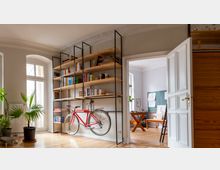 Heller Wohnraum mit Holzdielen, hohen Fenstern und Stuckdecke, dominiert von einem großen Regal aus Holz und schwarzem Metall mit Büchern und einem darin aufgehängten roten Fahrrad. Rechts führt eine offene weiße Flügeltür in ein weiteres Zimmer mit Arbeitstisch und Pinnwand.