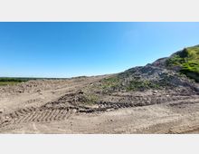 Ein Gelände mit aufgeschütteten Erdhügeln, teils mit spärlichem Grasbewuchs, unter klarem, blauem Himmel. Im Hintergrund erstreckt sich eine flache Landschaft mit vereinzelten Bäumen.