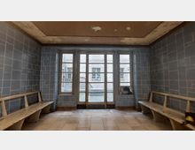 Ein schlichtes, alt wirkendes Wartezimmer mit blau gefliesten Wänden und zwei langen hölzernen Sitzbänken an den Seiten. Eine große Fensterfront mit mehreren Glasabschnitten führt zu einem gepflasterten Innenhof; an der Decke ist der Putz teilweise abgeblättert.