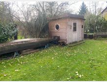 Holzschuppen mit rundem Fenster und kleiner Eingangstür auf einer Wiese, daneben ein umgedreht gelagertes Boot auf Stützen. Im Hintergrund befinden sich Bäume, Büsche und eine Holzbank.
