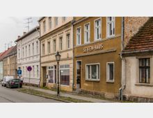 Eine schmale Straße mit historischen Wohnhäusern, darunter ein Gebäude mit der Aufschrift „SEIFEN-HAUS“. Die Fassaden variieren in Farben und Baustilen, und eine alte Straßenlaterne steht am Gehweg. Autos sind am Straßenrand geparkt.
