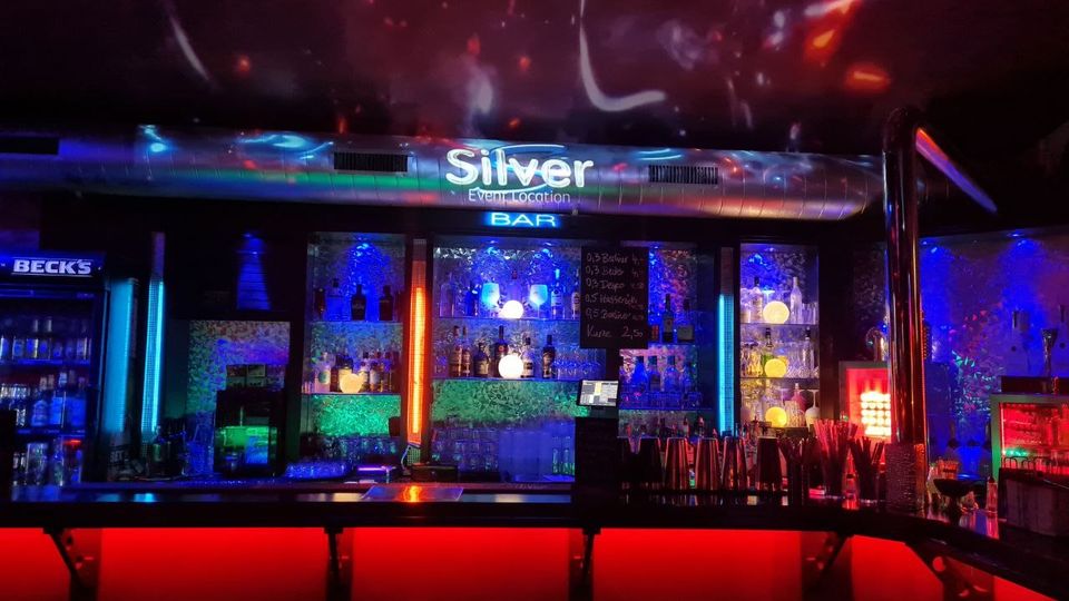 Innenansicht einer Bar mit beleuchteter Theke und Regalen voller Flaschen und Gläser, dominiert von blauem und rotem Neonlicht. Über der Bar hängt ein leuchtendes Schild „Silver Event Location“, an der Decke sind farbige Lichtprojektionen zu sehen.