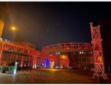 Industrielle Außenansicht bei Nacht mit roten, beleuchteten Stahlkonstruktionen, die als Brücken zwischen Backsteingebäuden dienen. Kopfsteinpflaster am Boden, umgeben von historischen Industriegebäuden mit großen Fenstern.