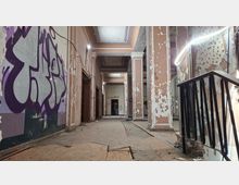 Verlassener Innenflur eines alten Gebäudes mit hohen Decken, Oberlichtern und Säulen; die Wände sind stark abgeblättert und teilweise mit Graffiti bedeckt. Im Vordergrund liegen beschädigte Bodenplatten und Schutt, rechts ist ein Treppengeländer zu sehen.