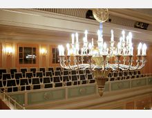 Ein prunkvoller Kronleuchter mit mehreren Kerzenlampen hängt in einem klassischen Theater- oder Konzertsaal. Im Hintergrund sind Sitzreihen auf einer Galerie sowie Wände mit verzierter Holzvertäfelung und warm beleuchteten Wandleuchten zu sehen.