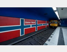 U-Bahn-Station mit dem Namen „Osloer Straße“ auf einer Wand in den Farben der norwegischen Flagge. Ein gelber U-Bahn-Zug fährt auf einem Gleis ein, der Boden besteht aus hellen Fliesen mit dunklen Streifen.