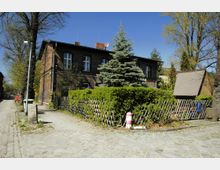 Backsteinhaus mit weißen Fenstern, umgeben von einer Hecke und einem schrägen Holzzaun, neben einem kleinen Straßenzug mit Kopfsteinpflaster. Im Vordergrund stehen eine Laterne und ein rot-weißes Poller, im Hintergrund sind Bäume und ein kleines Gartengebäude zu sehen.