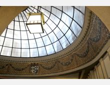 Ein dekoratives Glasdach mit einer runden Kuppelstruktur, unterteilt in geometrische Glasflächen, wird von einem Hängeleuchter beleuchtet. Darunter befindet sich eine verziertes Mosaikfries mit Wappen und ornamentalem Dekor.