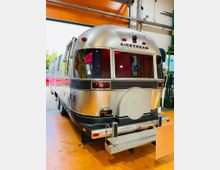Silberner Airstream-Wohnanhänger von hinten in einer Werkstatt- oder Garagenhalle mit orangefarbenem Rolltor, sichtbaren Leitungen und Regalen. Am Heck sind große Fenster, Rückleuchten, eine Reserveradabdeckung und eine ausklappbare Trittstufe zu sehen.