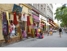 Straßenszene mit kleinen Geschäften in einer Altbauumgebung, darunter Boutiquen mit bunter Kleidung, Stoffen und Accessoires, die teils draußen ausgestellt sind. Auf dem Gehweg gehen Fußgänger an Schaufenstern und Verkaufsständen vorbei, während Bäume den Bürgersteig säumen.
