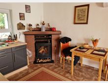 Ein gemütlicher Innenraum mit Kaminofen aus schwarzem Metall, eingebaut in eine Ziegelmauer. Daneben stehen eine Holztafel mit zwei Stühlen sowie eine kleine Küchenecke mit grauer Arbeitsfläche und Schränken. Decor-Elemente wie Bilder, ein Weidenkorb mit Karotten und andere Kleinigkeiten schmücken den Raum.