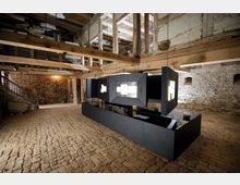 Ein historischer Innenraum mit freiliegendem Mauerwerk und Holzbalken. Im Zentrum befindet sich eine moderne, schwarz-graue Installation mit leuchtenden Aussparungen, umgeben von einem Boden aus unregelmäßigen Ziegelsteinen.