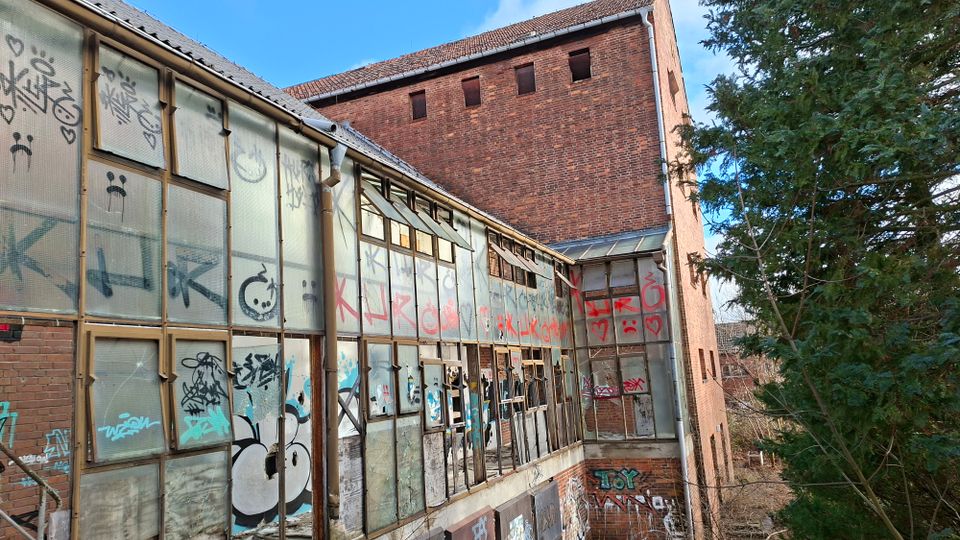 Verlassener Industrie- oder Lagerbau aus rotem Backstein mit einem langen Anbau aus Glas- und Metallrahmen, dessen Scheiben teils beschädigt und mit Graffiti bedeckt sind. Im Hintergrund ragt ein hoher Backsteinturm mit kleinen Fensteröffnungen auf, daneben stehen dichte Bäume am Rand des Geländes.