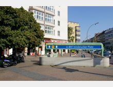 Eingang zur U-Bahn-Station „U-Bhf Innsbrucker Platz“ in einer städtischen Umgebung mit Wohn- und Geschäftshäusern im Hintergrund, darunter eine Apotheke. Im Vordergrund ein großer Baum, Fahrräder und ein Roller auf einem gepflasterten Gehweg, rechts vorbeifahrende Autos auf der Straße.