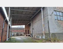 Überdachte Ladezone eines Industriegebäudes mit hohem Metallvordach auf Stahlstützen und Betonboden zwischen Ziegel- und Betonwänden. Im Hintergrund sind weitere niedrige Fabrikgebäude mit Fensterreihen und teils zugemauerten Öffnungen zu sehen.