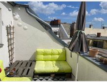Kleine Dachterrasse mit hellen Wänden, gelben Paletten-Sitzmöbeln und einem zusammengefalteten Sonnenschirm. Im Hintergrund sind umliegende Dächer und der blaue Himmel mit vereinzelten Wolken zu sehen.