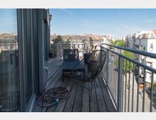 Ein Balkon mit Holzboden und Metallgeländer, ausgestattet mit einem schwarzen Stuhl, einer Liege und Pflanzen in Töpfen. Im Hintergrund ist eine ruhige Stadtansicht mit Wohnhäusern und Bäumen unter einem klaren blauen Himmel zu sehen.