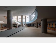 Großer, moderner Innenraum eines Gebäudes mit Betonpfeilern, geschwungenen Wänden und einer Glasfassade, die viel Tageslicht hereinlässt. Eine breite Treppe mit Holzgeländer führt zu einer oberen Etage, und im Hintergrund befinden sich eine Empfangstheke sowie Holzelemente an den Wänden.