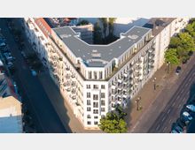 Wohngebäude in Ecklage mit heller Fassade und zahlreichen Balkons, umgeben von zwei Straßen mit parkenden Autos und vereinzelten Bäumen. Auf dem flachen Dach des Gebäudes befinden sich technische Anlagen und begrünte Bereiche.