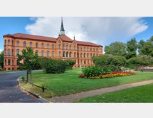 Historisches Backsteingebäude mit zahlreichen großen Fenstern und einem kleinen Uhrenturm, umgeben von einer gepflegten Parkanlage mit grünen Rasenflächen, Blumenbeeten und Bäumen sowie geschwungenen Wegen.