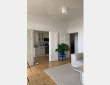 Bright, minimalist living room with white paneled double doors opening into a small white kitchen with tile backsplash and light wood floors. A large circular ceiling light, a gray area rug, and a potted plant on a blue stool sit near a doorway leading to another room.