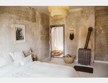 Schlafzimmer mit einem schlichten Bett, weißen Bettbezügen und rauen, unverputzten Wänden. Neben einem Kaminofen steht ein Korb mit Holz, und durch eine Tür ist ein Raum mit hellem Vorhang und Teppich zu sehen.