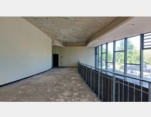 Ein leerer Innenraum mit abblätternder Decke und beschädigtem Boden. Auf der rechten Seite befinden sich bodentiefe Fenster mit Blick auf eine grüne Außengegend, links eine schlichte, helle Wand mit einer Tür.