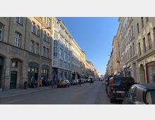 Straßenszene in einer europäischen Stadt mit Reihen historischer Wohn- und Geschäftshäuser aus hellen und Backsteinfassaden, flankiert von parkenden Autos. Im Vordergrund sind Fußgänger, Radfahrer und Cafés zu sehen, während die Straße von Sonnenlicht beleuchtet wird.