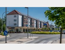 Mehrstöckiges Wohn- und Geschäftsgebäude mit weißer Fassade, roten Balkonen und Solaranlagen auf dem Dach; im Vordergrund ein Kreisverkehr mit bepflanzter Insel und einer modernen Skulptur, umgeben von Gehwegen und Bäumen.