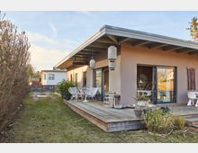 Flacher Bungalow mit überdachter Terrasse, Holzboden und modernen Hängeleuchten, umgeben von einem Garten mit Rasenfläche und seitlicher Hecke; die Terrasse ist mit weißen Stühlen und Pflanzen dekoriert. Im Hintergrund führt ein Weg zu einem geschlossenen Metalltor.
