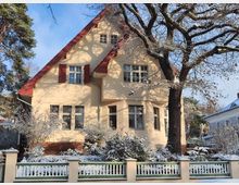 Gelbes Einfamilienhaus mit rotem Satteldach und weißen Fenstern, umgeben von einem schneebedeckten Garten mit Bäumen und Sträuchern. Im Vordergrund steht ein Zaun mit weißen Latten und grünen Details, daneben teils blauer Himmel und umliegende Gebäude.