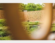 Moderner Garten mit symmetrisch angeordneten Beeten, niedrigen Hecken und einem rechteckigen Wasserbecken, eingefasst von einer dekorativen Wand aus durchbrochenen, rötlichen Metallplatten mit eingearbeiteten Schriftmustern; im Hintergrund dichte grüne Vegetation.