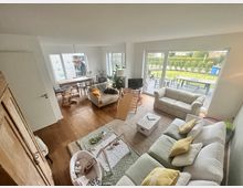 Wohnzimmer eines modernen Hauses mit Parkettboden, einer weißen Couchgarnitur und einem kleinen Esstisch mit Stühlen in einer Ecke. Große Fenster lassen viel Tageslicht herein und führen zu einem Garten mit Terrasse und Sitzmöbeln. Im Raum befinden sich auch Spielzeuge und Dekorationen für ein Baby.