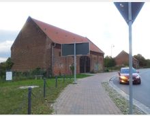 Backsteingebäude mit rotem Ziegeldach neben einer gepflasterten Straße, vermutlich eine alte Scheune oder Lagerhalle. Im Vordergrund ein kleiner Grünstreifen mit Kettengeländer, Verkehrsschildern und einem parkenden Auto am Straßenrand.