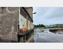 Industriegebäude mit verwitterten, rostigen Metalltoren auf einer erhöhten Rampe. Der umliegende Hof besteht aus Betonflächen mit vereinzeltem Grasbewuchs und Pfützen, im Hintergrund sind weitere Gebäude und Bäume zu sehen.