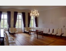 Ein heller Raum mit großen Fenstern, die von dunkelvioletten Vorhängen eingerahmt sind, und einem zentralen Kronleuchter. Der Raum ist mit einer langen Reihe gepolsterter Stühle und einem Holztisch auf einem glänzenden Holzboden möbliert.